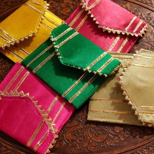 Op de afbeelding: Een verzameling van vijf rechthoekige stoffen enveloppen in geel, felroze, groen en gedempt goud. Elke envelop heeft een puntige flap, metallic gouden strepen en een decoratieve gouden schulprand langs de randen.