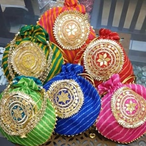 Op de afbeelding: Een verzameling van zes buideltjes met trekkoord in diverse kleuren zoals groen, blauw, rood, oranje en roze, elk voorzien van een gestreept stoffen patroon en een grote, ronde, goudkleurige decoratieve medaillon met een stervormige spiegel.