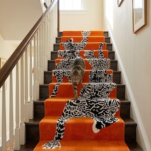 May include: A staircase runner featuring a vibrant orange carpet with a repeating black and white leopard print pattern. A brown tabby cat is walking down the center of the stairs, positioned on the patterned carpet runner between the wooden railings.
