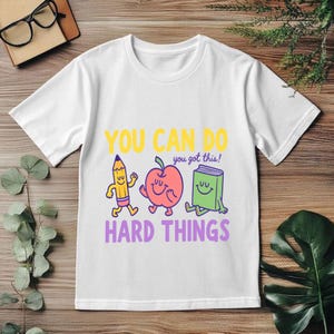 May include: A white short-sleeve t-shirt featuring a colorful graphic of a smiling pencil, apple, and book. The text reads YOU CAN DO you got this! HARD THINGS in yellow, purple, and blue, with the characters outlined in dark blue ink.