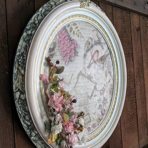 May include: A white oval frame with a green and gold trim. The frame contains a vintage floral print with pink flowers and birds. A bouquet of pink and white flowers is attached to the frame.