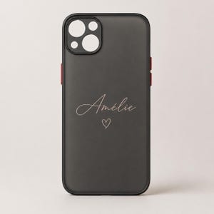 Peut inclure: Une coque de téléphone noir mat avec une découpe pour l'appareil photo en haut à gauche. Le centre de la coque affiche le prénom Amélie écrit en écriture cursive fine et claire, placé juste au-dessus d'un petit contour de cœur dessiné à la main.