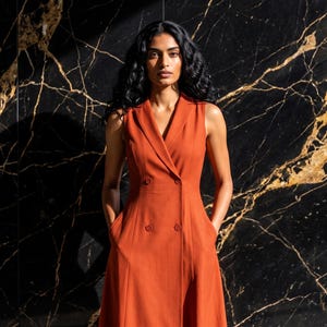 May include: A sleeveless, burnt orange midi dress featuring a double-breasted front with four matching buttons, a notched lapel collar, and two side pockets. The garment has a tailored bodice that transitions into a flared skirt silhouette.