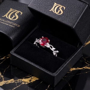 May include: A silver ring featuring a round, deep red gemstone center stone flanked by small clear stones, set in a vine-like band. The ring is displayed inside an open black textured box with the gold logo text JEWELSGALLERYSTUDIOS printed on it.