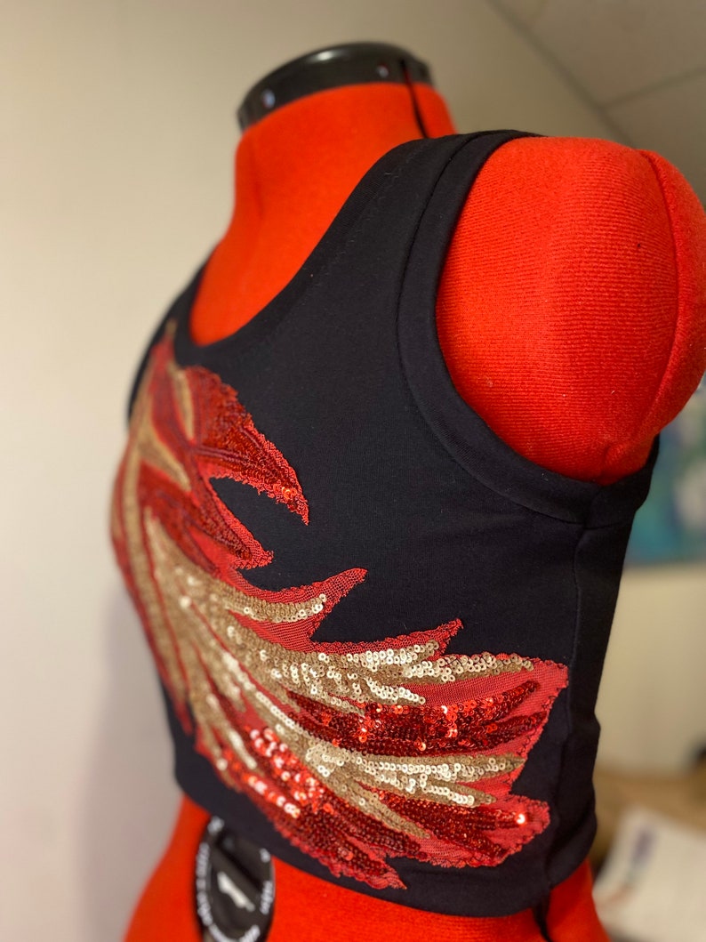 May include: A black tank top with a red and gold sequined wing design. The tank top is cropped and has a sleeveless design.