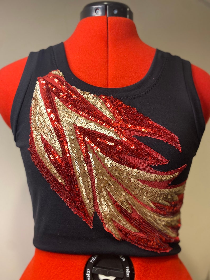 May include: A black tank top with a red and gold sequined design on the front. The design is a stylized feather shape.