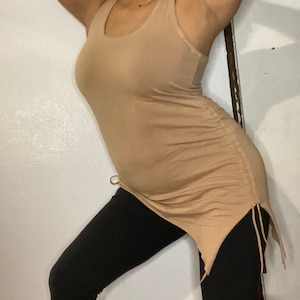 May include: A woman wearing a light brown tank top with a side tie detail and black leggings.