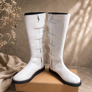 May include: A pair of knee-high white boots featuring a flat black sole, a vertical zipper along the inner side, and four horizontal straps with snap closures on the outer side. The boots have a smooth finish and a rounded toe design.