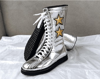 Puede incluir: Un par de botas de media caña con acabado plateado metálico, cordones blancos, suela negra con relieve y dos parches de estrellas doradas con bordes negros en el lado exterior de la caña. Las botas tienen una puntera redondeada.