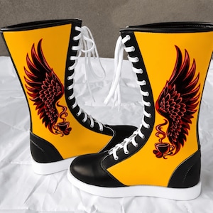 Puede incluir: Un par de botas acordonadas a media pantorrilla con exterior amarillo vibrante, ribetes negros y cordones blancos. Cada bota muestra un gráfico de una taza de café humeante con grandes alas emplumadas en rojo oscuro y negro.