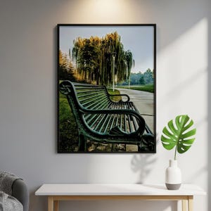 May include: A framed wall art print featuring a dark green metal park bench positioned on a paved path next to a large weeping willow tree. The scene captures a quiet outdoor setting with green grass and trees under a clear, bright sky.