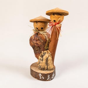 May include: A wooden figurine featuring two figures with conical hats, straw-like neck accents, and painted facial features. The figures are mounted on a circular base with white Japanese characters painted on the front edge of the wood.