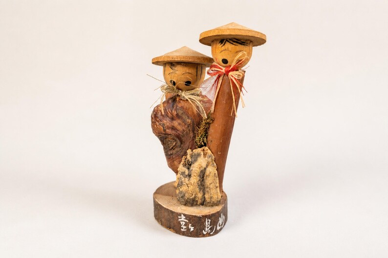 May include: A wooden figurine featuring two figures with conical hats, straw-like neck accents, and painted facial features. The figures are mounted on a circular base with white Japanese characters painted on the front edge of the wood.