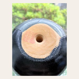K&ouml;nnte beinhalten: Ein schwarzes, abgerundetes Keramikobjekt mit glatter, dunkler Oberfl&auml;che. Der Boden weist eine kreisf&ouml;rmige, unglasierte, hellbraune Mitte mit einem zentralen Loch und einem kleinen, quadratischen roten Stempelabdruck auf der hellen Unterseite auf.