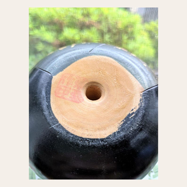 K&ouml;nnte beinhalten: Ein schwarzes, abgerundetes Keramikobjekt mit glatter, dunkler Oberfl&auml;che. Der Boden weist eine kreisf&ouml;rmige, unglasierte, hellbraune Mitte mit einem zentralen Loch und einem kleinen, quadratischen roten Stempelabdruck auf der hellen Unterseite auf.