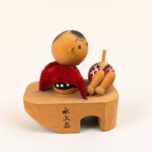 May include: A wooden kokeshi-style figurine featuring a round head with black hair, a red velvet-like fabric accent, and a separate small round object with a peg. The figure sits on a light brown wooden base with black Japanese characters printed on it.