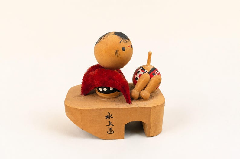 May include: A wooden kokeshi-style figurine featuring a round head with black hair, a red velvet-like fabric accent, and a separate small round object with a peg. The figure sits on a light brown wooden base with black Japanese characters printed on it.