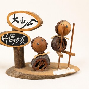 May include: A decorative figurine made of natural nuts and wood, featuring two small figures. One figure stands while the other uses wooden stilts. Two oval wooden signs are attached to a post, displaying the Japanese text 大山崎 and 竹馬坂.