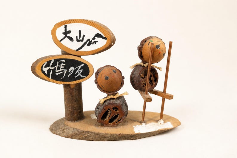May include: A decorative figurine made of natural nuts and wood, featuring two small figures. One figure stands while the other uses wooden stilts. Two oval wooden signs are attached to a post, displaying the Japanese text 大山崎 and 竹馬坂.