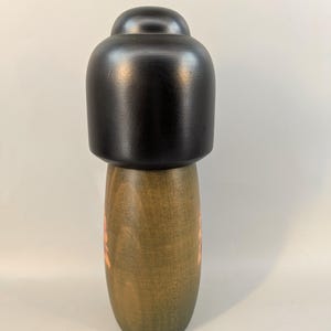 May include: A wooden pepper mill featuring a smooth, matte black top section and a cylindrical base with a natural olive-green wood grain finish. The base displays a subtle, light-colored floral pattern etched into the wood surface.