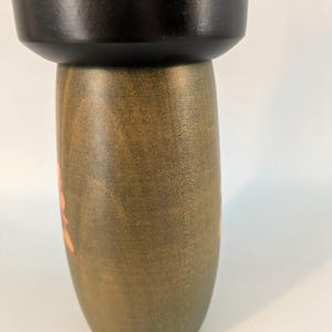 May include: A wooden kokeshi doll featuring a smooth, matte black cylindrical head section resting atop a rounded, olive-green body. The body displays a visible wood grain texture and a faint, light-colored floral pattern painted on the side.