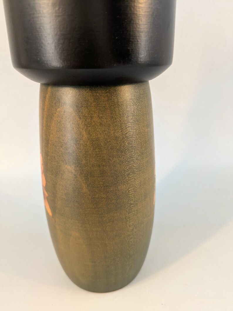 May include: A wooden kokeshi doll featuring a smooth, matte black cylindrical head section resting atop a rounded, olive-green body. The body displays a visible wood grain texture and a faint, light-colored floral pattern painted on the side.