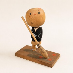 May include: A wooden kokeshi doll with a large round head, a black jacket, and a tan skirt. The doll holds a long wooden broom and stands on a rectangular wooden base decorated with small red painted star shapes and a dark grey rectangular patch.