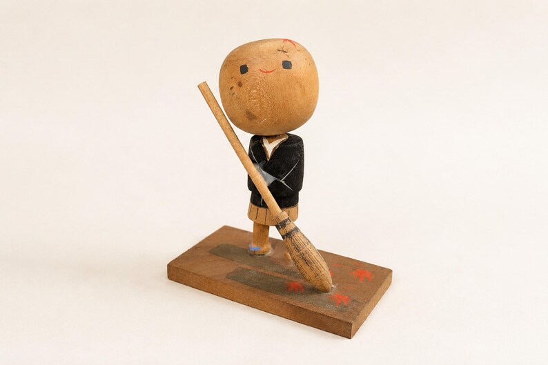 May include: A wooden kokeshi doll with a large round head, a black jacket, and a tan skirt. The doll holds a long wooden broom and stands on a rectangular wooden base decorated with small red painted star shapes and a dark grey rectangular patch.
