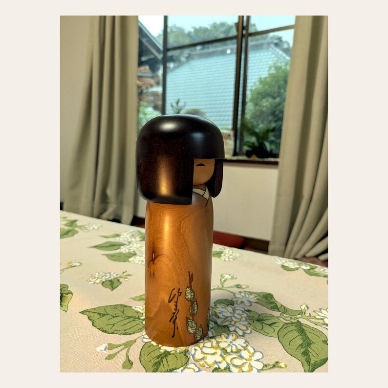 K&ouml;nnte beinhalten: Eine h&ouml;lzerne Kokeshi-Puppe mit einem glatten, dunkelbraunen, abgerundeten Kopf und einem zylindrischen K&ouml;rper aus hellbraunem Holz. Der K&ouml;rper zeigt ein handgemaltes gr&uuml;nes Pflanzenmotiv und schwarze japanische Schriftzeichen an der Basis.