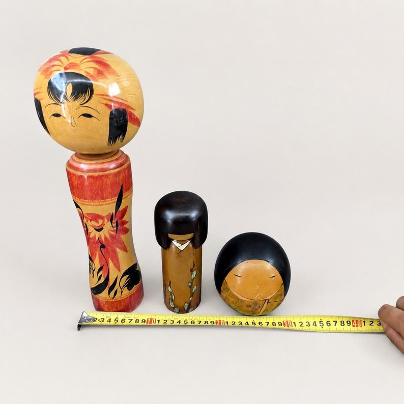 K&ouml;nnte beinhalten: Drei h&ouml;lzerne Kokeshi-Puppen in verschiedenen Gr&ouml;&szlig;en. Die gr&ouml;&szlig;te Puppe hat einen runden Kopf und rote Blumenmuster. Ein gelbes Ma&szlig;band liegt davor und zeigt, dass die gr&ouml;&szlig;te Puppe eine H&ouml;he von etwa 18 cm hat und die anderen kleiner sind.