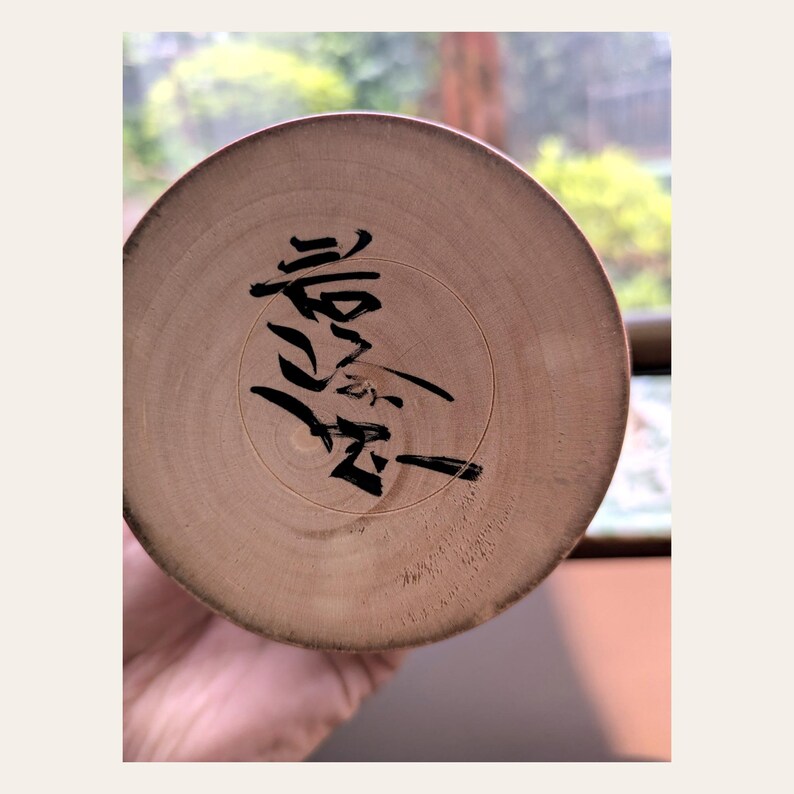 K&ouml;nnte beinhalten: Ein kreisf&ouml;rmiger, heller Holzsockel mit einer markanten schwarzen Tintenkalligrafie-Signatur in der Mitte der Oberfl&auml;che. Die Holzmaserung ist sichtbar und zeigt nat&uuml;rliche konzentrische Ringe sowie eine glatte, flache Textur.