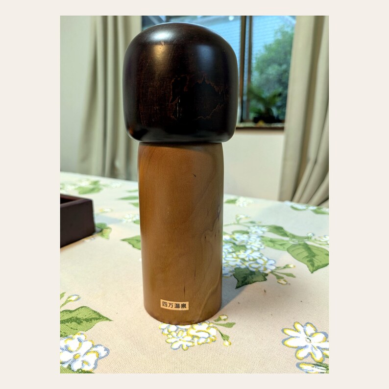 K&ouml;nnte beinhalten: Eine h&ouml;lzerne Kokeshi-Puppe mit einem glatten, dunkelbraunen, abgerundeten Kopf und einem zylindrischen, hellbraunen K&ouml;rper. Ein kleiner rechteckiger Aufkleber mit dem japanischen Text 四万温泉 ist am unteren vorderen Teil des hellen Holzsockels angebracht.