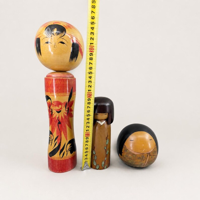 K&ouml;nnte beinhalten: Drei h&ouml;lzerne Kokeshi-Puppen unterschiedlicher Gr&ouml;&szlig;e neben einem gelben Ma&szlig;band. Die gr&ouml;&szlig;te Puppe hat einen runden Kopf und einen roten K&ouml;rper mit Blumenmuster und misst 29 cm. Eine kleinere braune Puppe zeigt gr&uuml;ne Blattdetails und eine runde Puppe hat ein schwarzes Oberteil.