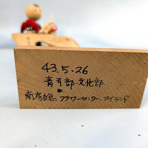 May include: A rectangular wooden block with black handwritten Japanese text that reads 43.5.26, 青年部・文化部, and 南房総フラワーセンター・アイランド. The block has a light brown wood grain finish and is positioned in front of a blurred wooden figurine.