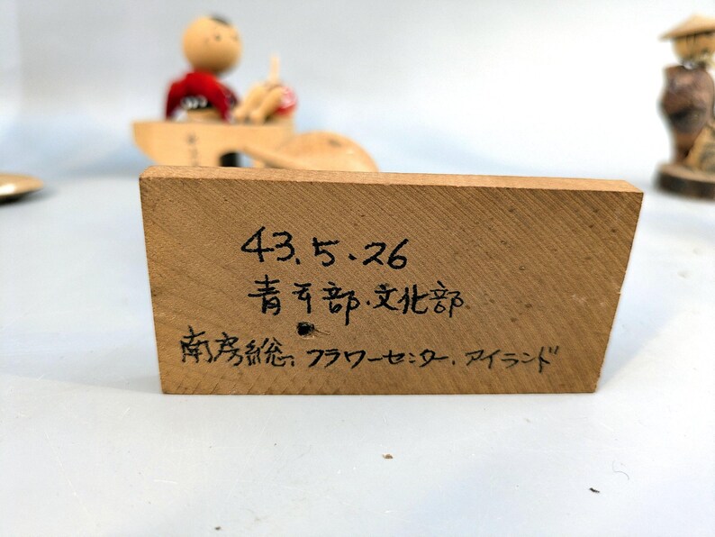 May include: A rectangular wooden block with black handwritten Japanese text that reads 43.5.26, 青年部・文化部, and 南房総フラワーセンター・アイランド. The block has a light brown wood grain finish and is positioned in front of a blurred wooden figurine.
