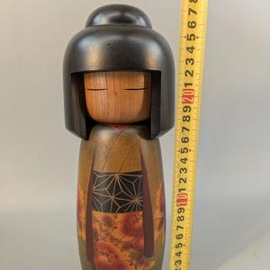May include: A wooden kokeshi doll with a smooth black rounded hairstyle and a natural wood face. The body features a dark green base with a black geometric patterned obi and orange floral designs. A yellow measuring tape shows a height of 23 cm.