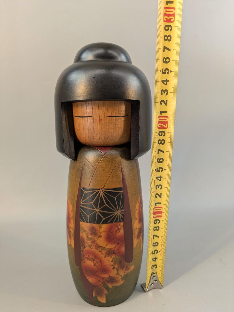 May include: A wooden kokeshi doll with a smooth black rounded hairstyle and a natural wood face. The body features a dark green base with a black geometric patterned obi and orange floral designs. A yellow measuring tape shows a height of 23 cm.