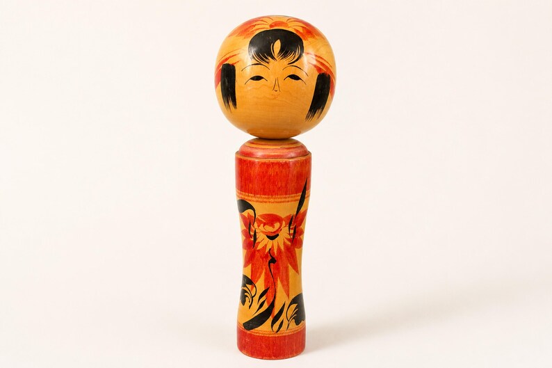 K&ouml;nnte beinhalten: Eine h&ouml;lzerne Kokeshi-Puppe mit einem kugelf&ouml;rmigen Kopf, bemaltem Gesicht, schwarzem Haar und orangefarbenen Blumenakzenten. Der zylindrische K&ouml;rper ist in Gelb- und Rott&ouml;nen mit schwarzen und orangefarbenen Blumenmustern und glatter Oberfl&auml;che.