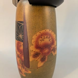 May include: A cylindrical wooden Kokeshi doll featuring a smooth olive green body decorated with painted orange and brown floral motifs. The doll has a solid black rounded head piece and a natural light wood tone visible at the neck area.
