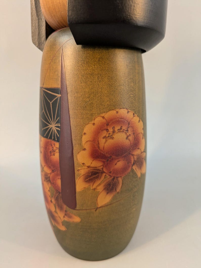 May include: A cylindrical wooden Kokeshi doll featuring a smooth olive green body decorated with painted orange and brown floral motifs. The doll has a solid black rounded head piece and a natural light wood tone visible at the neck area.