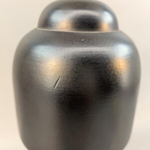 May include: A smooth, matte black, rounded wooden cap with a small, thin scratch on its surface. The cap features a domed top section that transitions into a wider, cylindrical base, showcasing a dark, uniform finish with subtle wood grain.