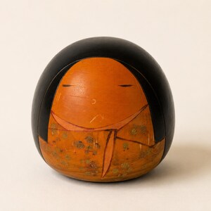K&ouml;nnte beinhalten: Eine abgerundete Holzfigur mit einem glatten schwarzen Oberteil und einem hellorangen Gesicht. Der untere Teil zeigt ein gemustertes orangefarbenes Kleidungsst&uuml;ck mit goldenen Blumenakzenten. Das Gesicht hat einfache, d&uuml;nne horizontale Linien als Augen.