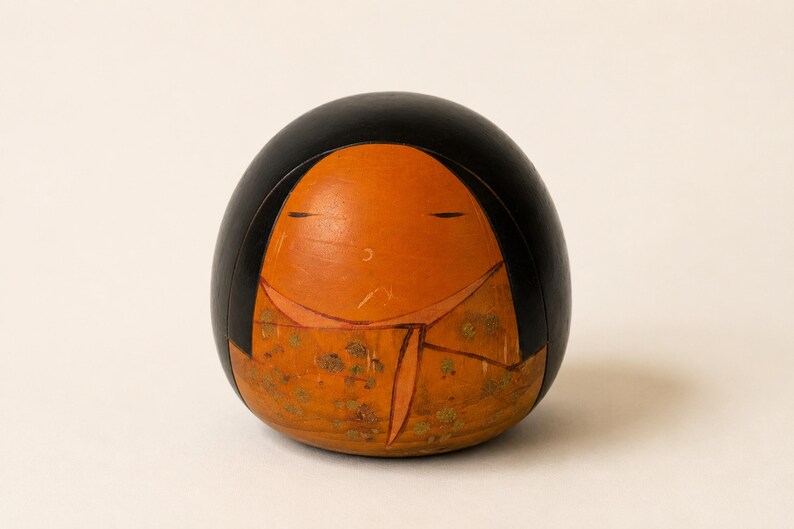 K&ouml;nnte beinhalten: Eine abgerundete Holzfigur mit einem glatten schwarzen Oberteil und einem hellorangen Gesicht. Der untere Teil zeigt ein gemustertes orangefarbenes Kleidungsst&uuml;ck mit goldenen Blumenakzenten. Das Gesicht hat einfache, d&uuml;nne horizontale Linien als Augen.