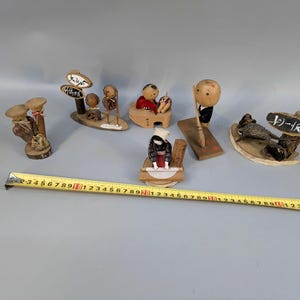 May include: A collection of six small wooden figurines with painted details, including figures with signs and tools, arranged alongside a yellow measuring tape that shows a length of approximately 55 cm across the display.