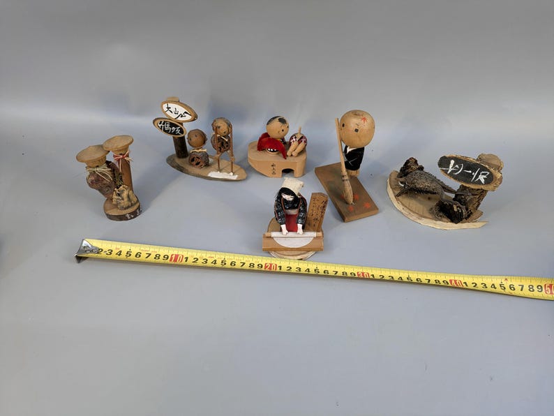 May include: A collection of six small wooden figurines with painted details, including figures with signs and tools, arranged alongside a yellow measuring tape that shows a length of approximately 55 cm across the display.