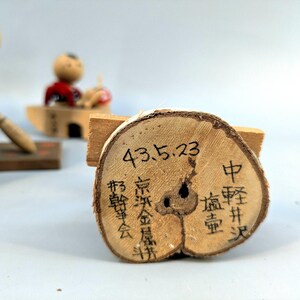May include: A circular wooden slice with bark edges featuring black ink calligraphy and the date 43.5.23. In the background, two small wooden figurines are visible, one holding a broom and another seated in a small wooden boat structure.