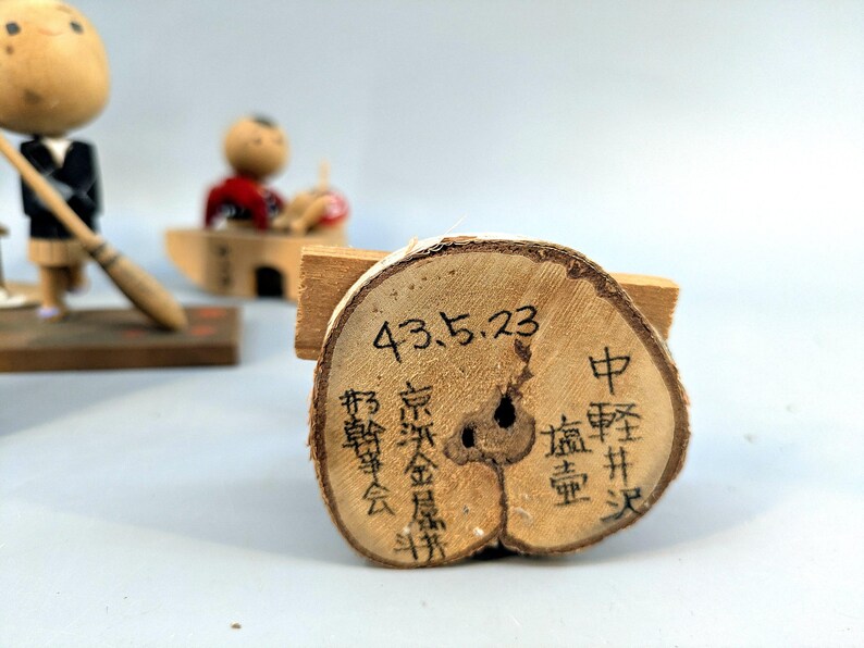 May include: A circular wooden slice with bark edges featuring black ink calligraphy and the date 43.5.23. In the background, two small wooden figurines are visible, one holding a broom and another seated in a small wooden boat structure.