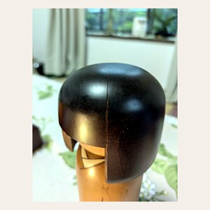 K&ouml;nnte beinhalten: Eine h&ouml;lzerne Kokeshi-Puppe mit einem glatten, dunkelbraunen, abgerundeten Kopf, der einen sichtbaren vertikalen Riss an der Seite aufweist. Der untere Teil der Puppe besteht aus hellerem Naturholz und zeigt ein einfaches Kragendetail am Hals.