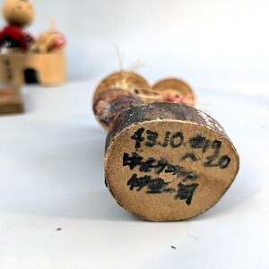 May include: A wooden kokeshi doll with a rounded head and a cylindrical body. The base of the doll features black handwritten Japanese text and numbers, including 43.10.19, indicating specific markings on the natural wood grain surface.