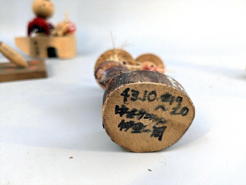 May include: A wooden kokeshi doll with a rounded head and a cylindrical body. The base of the doll features black handwritten Japanese text and numbers, including 43.10.19, indicating specific markings on the natural wood grain surface.